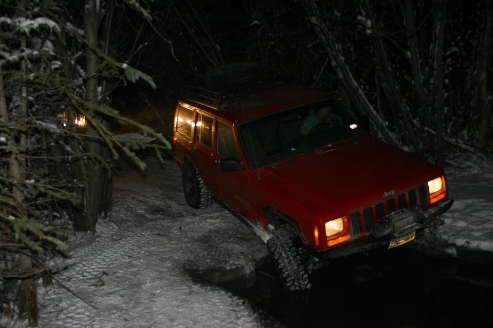 Red Jeep Cherokee XJ with a winch on the front bumper driving on a snowy, wooded road at night. A spare tire is mounted on the roof rack