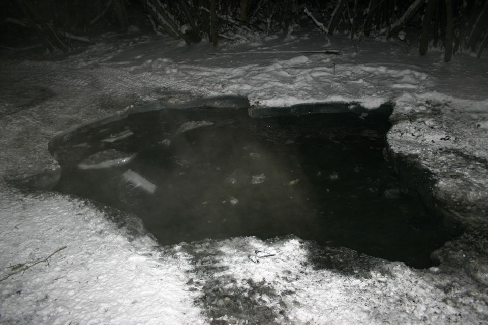 Hole in ice with dark water and broken ice chunks, surrounded by snow and tire tracks