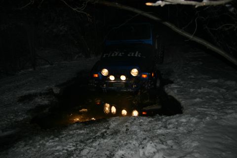 2003 Jeep TJ - A blue 2003 Jeep TJ, identified by its distinctive body style and front grille, is stuck in a muddy, snowy area at night, with "ride ak" vi…