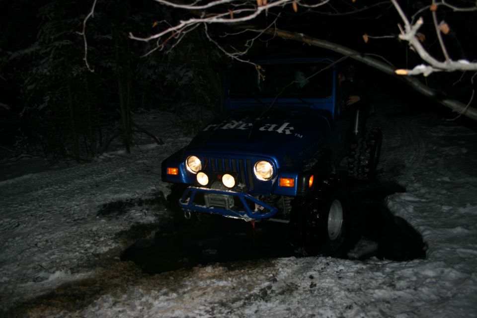 Blue Jeep Wrangler with "ride ak" text on hood, aftermarket bumper, and auxiliary lights, parked on a snowy path at night