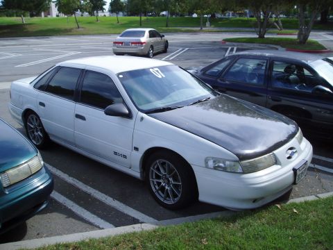 1993 Ford Taurus - A white sedan with a carbon fiber hood is parked in a lot, with other vehicles nearby.