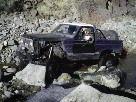 1986 Ford Bronco - A modified Ford Bronco traverses a rocky stream, showing significant damage to its front end.