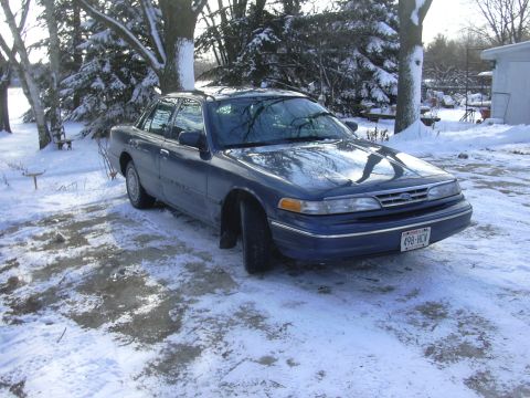 1996 Ford Crown Victoria - A blue sedan sits in a snow-covered yard with trees and a building in the background.
