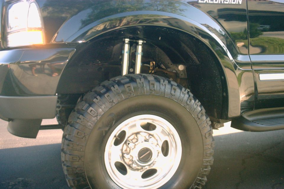Front wheel of lifted black Ford Excursion with mud-terrain tire and dual shocks visible in wheel well