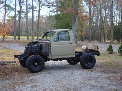 1991 Ford Bronco - A tan pickup truck chassis sits outdoors with its front end disassembled, revealing the engine.