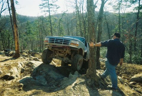1993 Ford Bronco - Man watches a 1993 Ford Bronco tackle a challenging rocky trail in a wooded area.