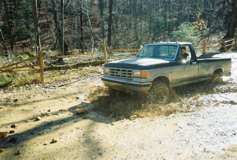 1993 Ford Bronco - A two-tone Ford F-Series truck navigates a muddy trail, kicking up water and dirt.