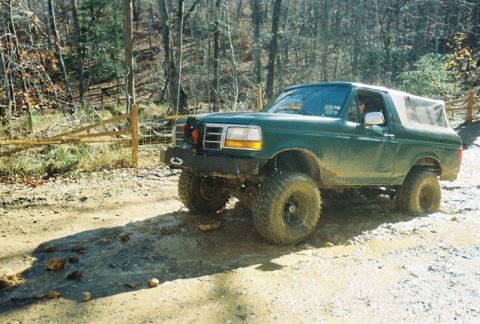 1993 Ford Bronco - A lifted green Ford Bronco navigates a muddy, wooded trail with a wooden fence in the background.