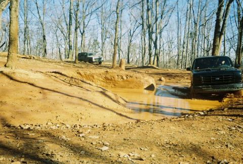 1993 Ford Bronco - Two SUVs navigate a muddy trail in a forest, one is a black Ford Bronco.