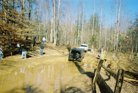 1993 Ford Bronco - Two SUVs, including a 1993 Ford Bronco, traverse a challenging muddy trail in a wooded area.
