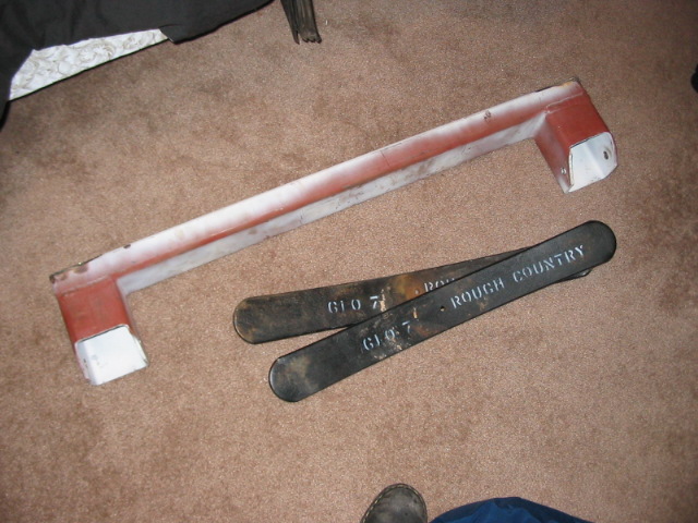 Two black metal bars with white lettering that says "ROUGH COUNTRY" and "GIO 7" sit on a carpet next to a red and white metal part