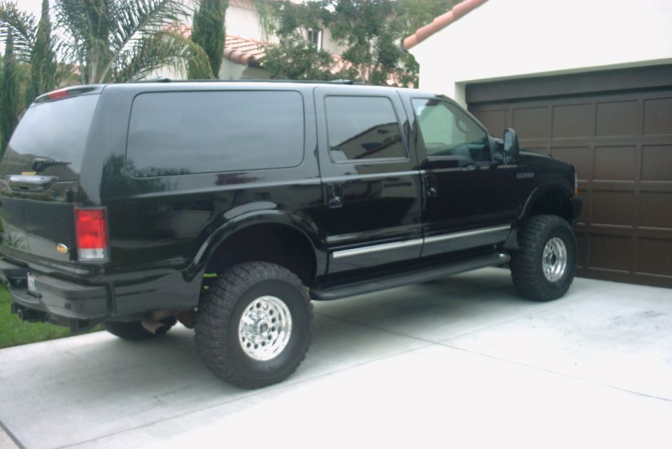 Black lifted Ford Excursion on big tires parked in a driveway beside a brown garage door and palms