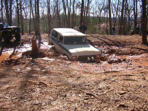 1979 Ford Bronco - A tan and blue Ford Bronco is stuck in a muddy rut in a wooded area, with another off-road vehicle visible to the left.