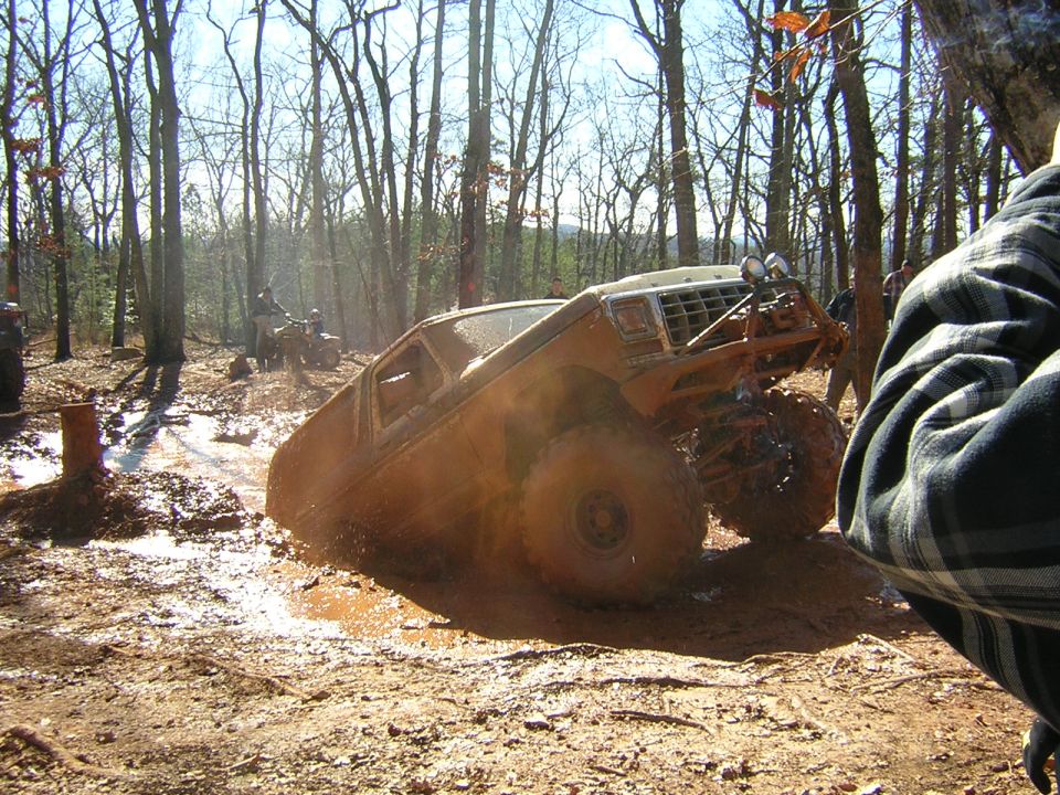 Orange Ford 4x4 climbing out of a muddy pit at an off-road event, front 3/4 view