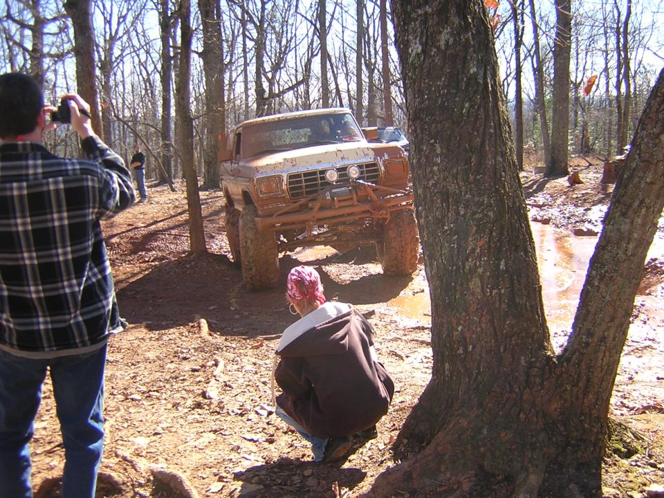 Muddy 1979 Ford Bronco with winch and auxiliary lights driving through a muddy trail in a forest