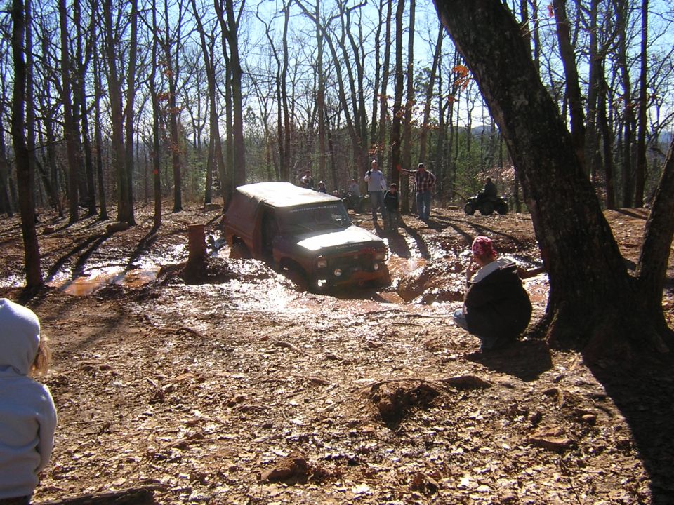 Green SUV crossing a muddy off-road trail in woods with spectators watching