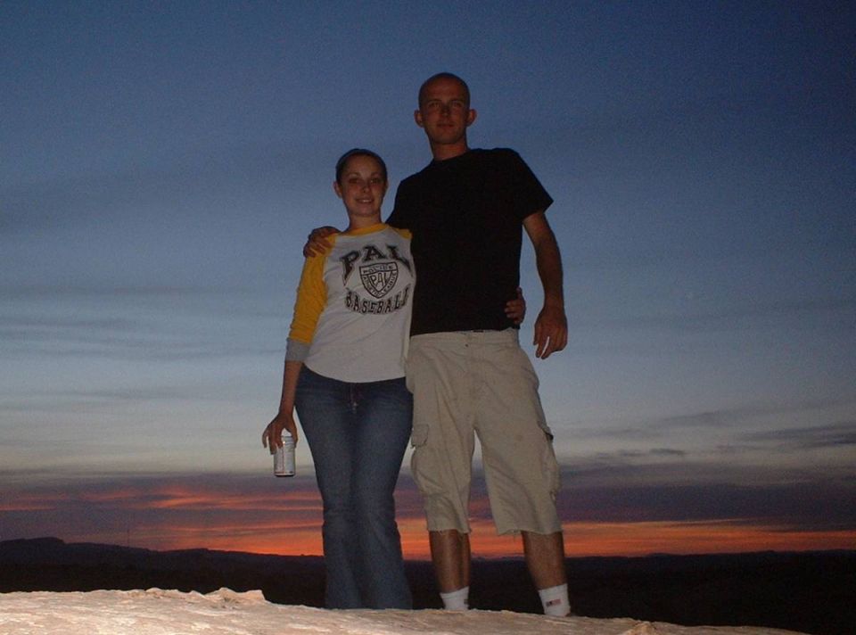 A man and woman posing together outdoors at dusk against an orange sunset sky.