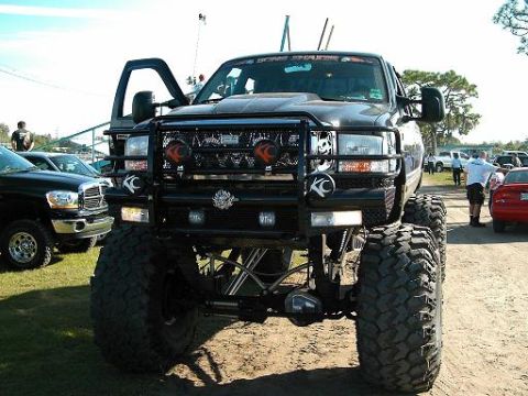 1986 Ford Bronco - A heavily modified black pickup truck with oversized tires and KC lights is parked outdoors, with other vehicles visible in the background.