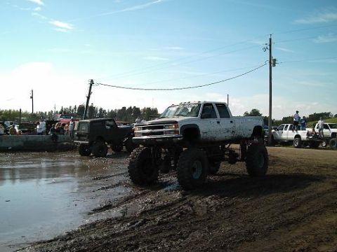 1986 Ford Bronco - Mud bogging event with multiple lifted trucks and SUVs in a muddy field under a blue sky.