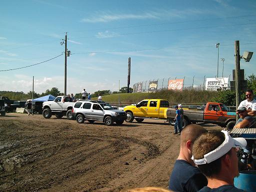 Several trucks and an SUV lined up on a dirt track, with spectators in the foreground and a race track in the background