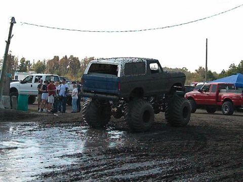 1986 Ford Bronco - Muddy 1986 Ford Bronco monster truck at an outdoor event with spectators and other vehicles.