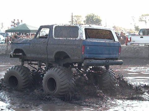 1986 Ford Bronco - This lifted Ford Bronco is covered in mud as it drives through a mud bog, with spectators in the background.