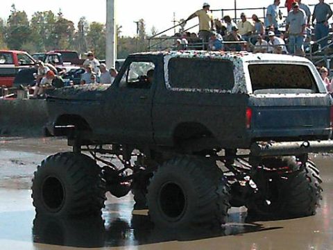 1986 Ford Bronco - A heavily modified 1980s Ford Bronco with oversized tires navigates a muddy track, with spectators in the background.