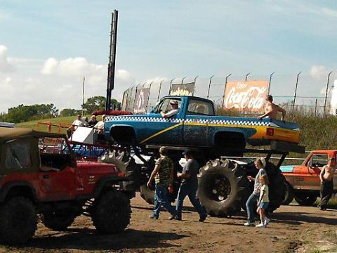 1986 Ford Bronco - A blue pickup truck with a yellow stripe and oversized tires is on display, with other vehicles and spectators present.