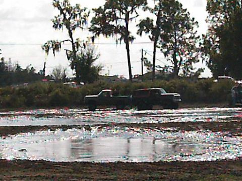 1986 Ford Bronco - Two trucks are in a muddy area with puddles and trees in the background.