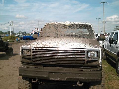 1986 Ford Bronco - A heavily muddied pickup truck sits parked at an outdoor event with other vehicles and spectators visible.