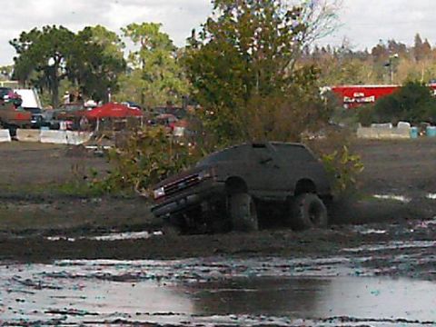 1986 Ford Bronco - A dark SUV navigates a muddy track with large tires, kicking up dirt.
