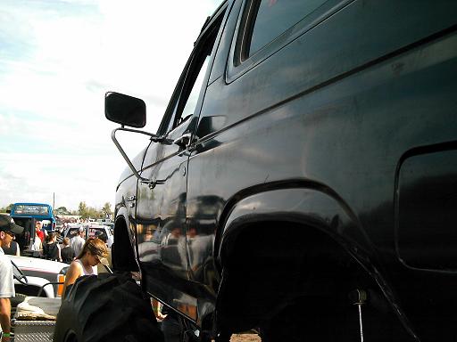 Dark SUV side view, lifted with large tires, in a lot with other vehicles and people