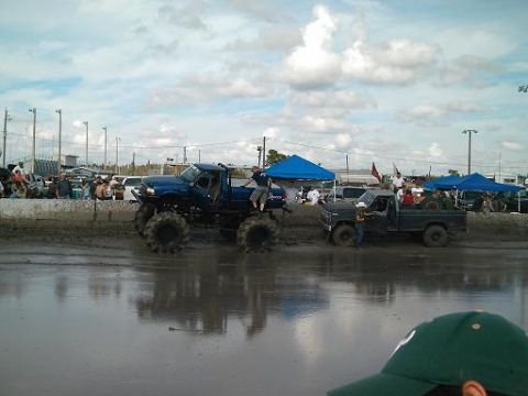 1986 Ford Bronco - Two trucks participate in a mud bog event with spectators watching from the sidelines.
