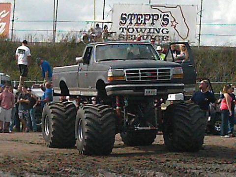 1986 Ford Bronco - Monster truck with massive tires sits in muddy terrain, front grille visible.