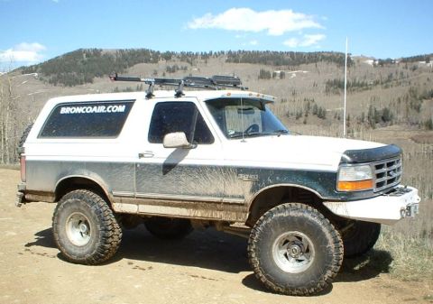 1993 Ford Bronco - White and gray two-tone Ford Bronco full-size SUV shown in 3/4 front view, lifted on large mud tires with steel wheels, roof rack, front br…