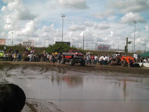 1986 Ford Bronco - A crowd watches as a lifted red truck and an orange tractor sit in a muddy field during an event.