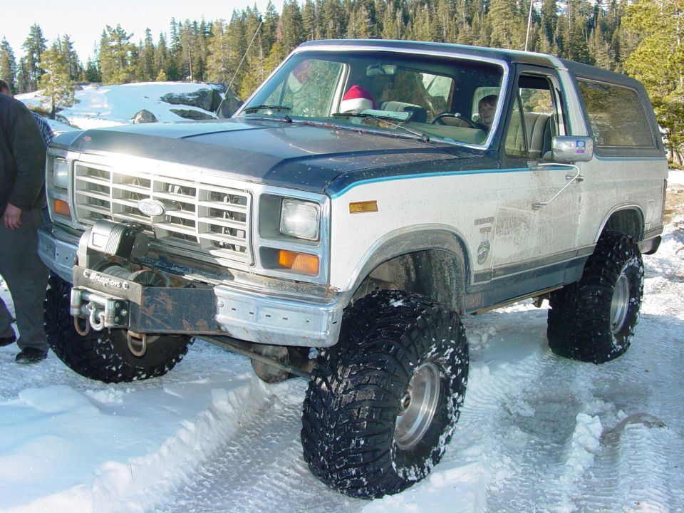 White and blue Ford Bronco with large tires and a winch, parked in snow