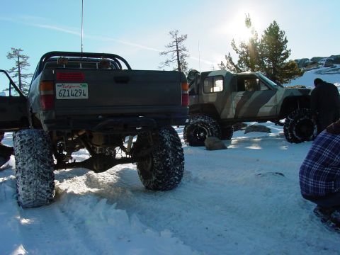 1986 Ford Bronco - Two heavily modified trucks with off-road tires sit in a snowy landscape under a bright sun.