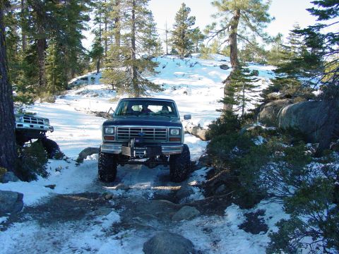1986 Ford Bronco - A lifted Ford Bronco navigates a snowy, rocky trail in a wooded area. Another vehicle is seen in the background.