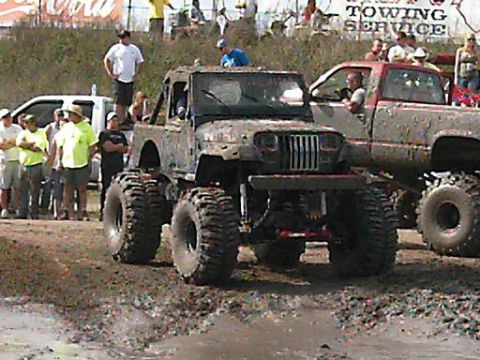 1986 Ford Bronco - An off-road vehicle navigates a muddy course, with a pickup truck visible to its right.