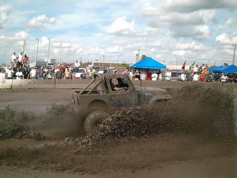 1986 Ford Bronco - A Jeep Wrangler churns up mud during a competition with onlookers in the background.