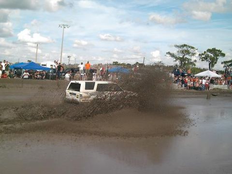 1986 Ford Bronco - A white SUV navigates a muddy track, sending up a large spray of mud as onlookers watch from the sidelines.