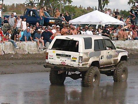 1986 Ford Bronco - A lifted white SUV navigates a muddy track with spectators watching from a concrete barrier.