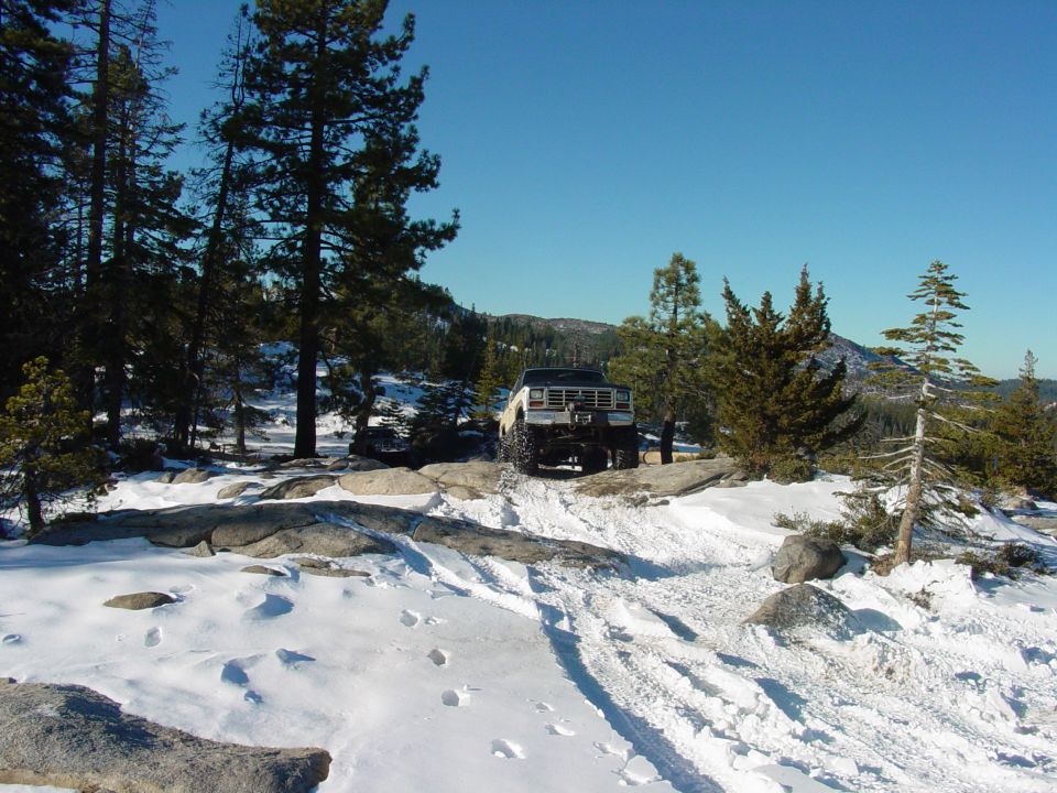 1986 Ford Bronco parked on a snowy trail with pine trees and mountains in the background