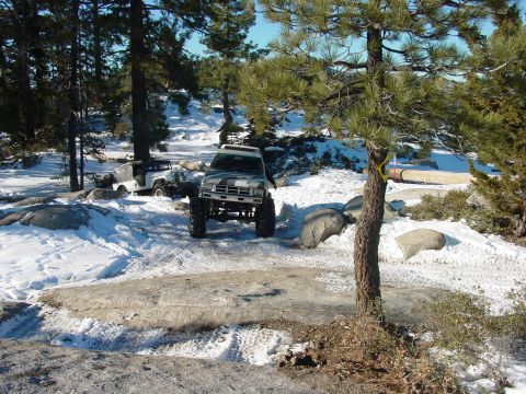 1986 Ford Bronco - Two off-road vehicles navigate a snowy trail in a wooded area.