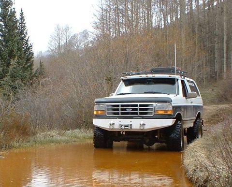 1993 Ford Bronco - A white two-tone Ford Bronco SUV with a black grille and hood, lift kit, oversized off-road tires, roof rack with light bar, and a whip ant…