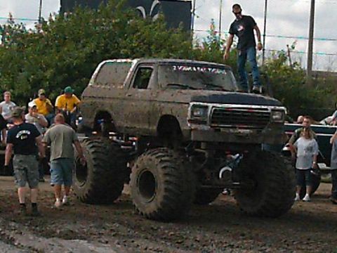 1986 Ford Bronco - A heavily modified truck with oversized tires and a lifted chassis is covered in mud, with spectators around.
