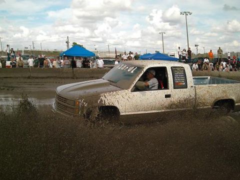 1986 Ford Bronco - Mud bogging event with a white pickup truck splashing through the mire.