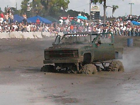 1986 Ford Bronco - A heavily modified truck navigates a muddy track during an event, with a large crowd watching from the sidelines.