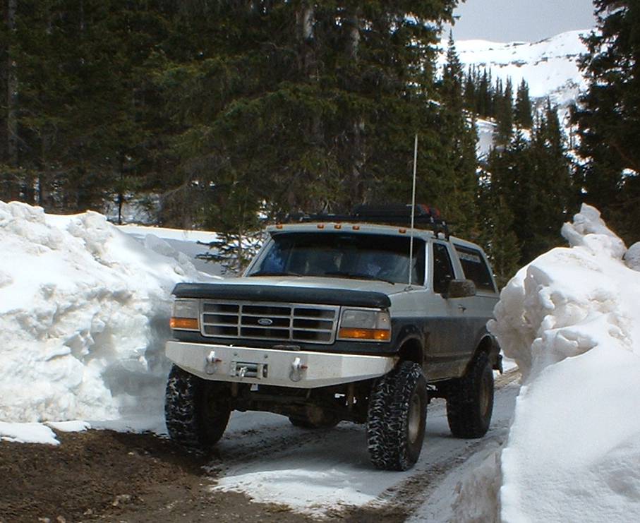 Lifted gray Ford Bronco with winch bumper on a snowy trail between deep snowbanks and pine trees